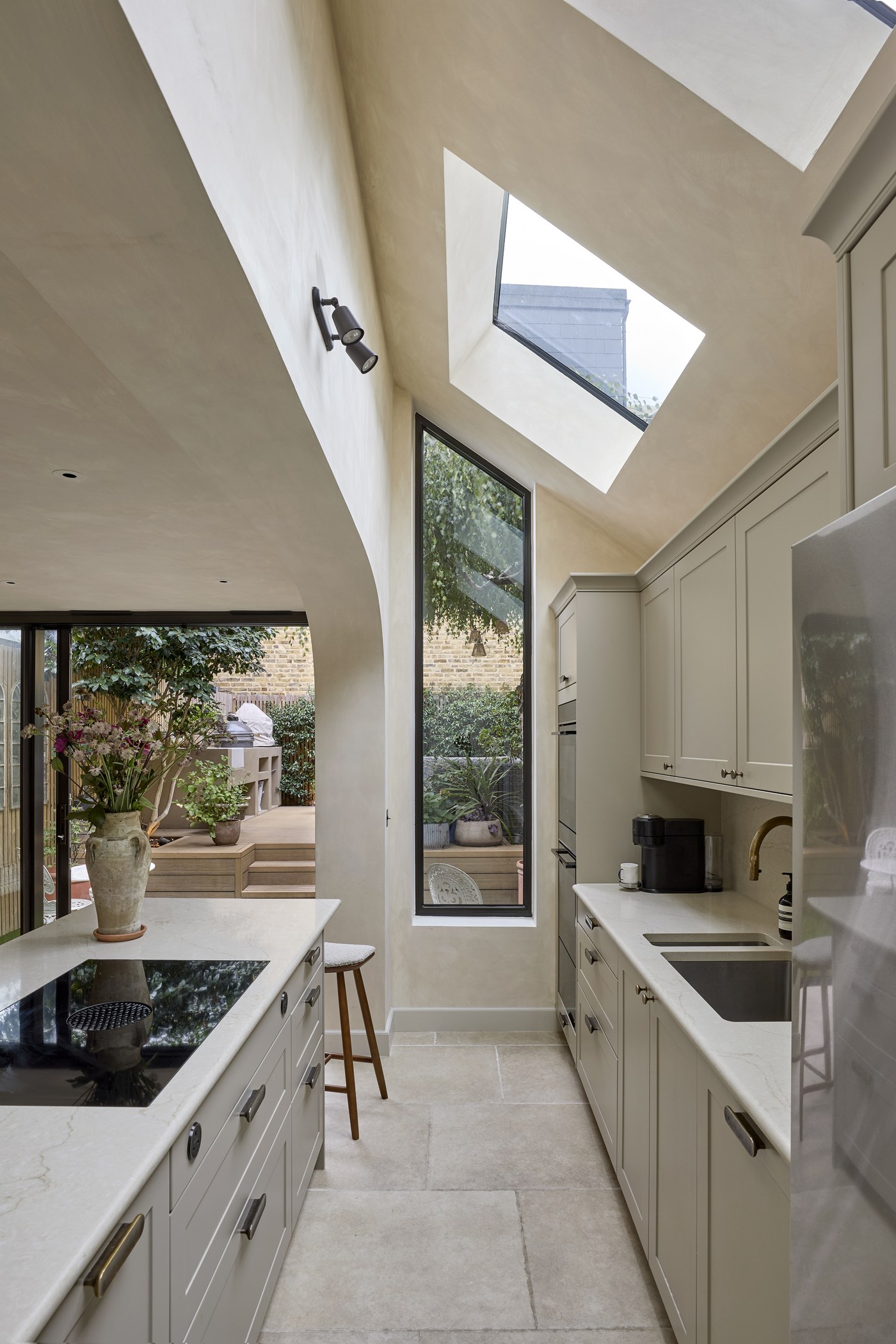 Honeywell Road SW11 — curved limewashed wall rising to a deep-set rooflight over the galley kitchen, with a full-height slot window to the garden and stone floor tiles